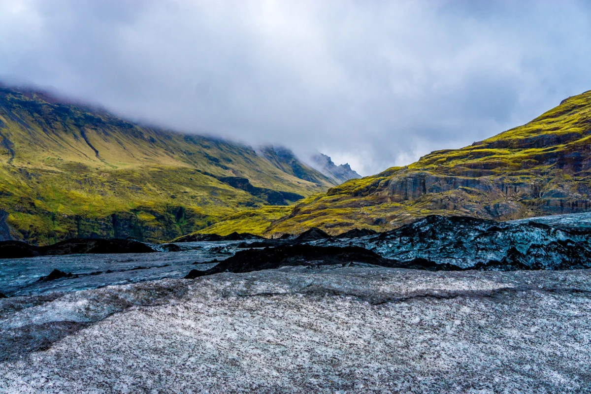 Dark ridges of glacier ice beneath green mountains and low clouds in Iceland